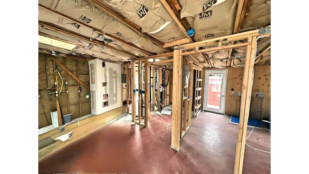 Interior of a home under renovation showing exposed wooden wall framing and ceiling joists. The space features copper plumbing pipes along the left wall, insulation visible in the exposed walls, and a reddish concrete floor. Natural light enters through a doorway in the background, and the room appears to be in the middle of construction or remodeling work.