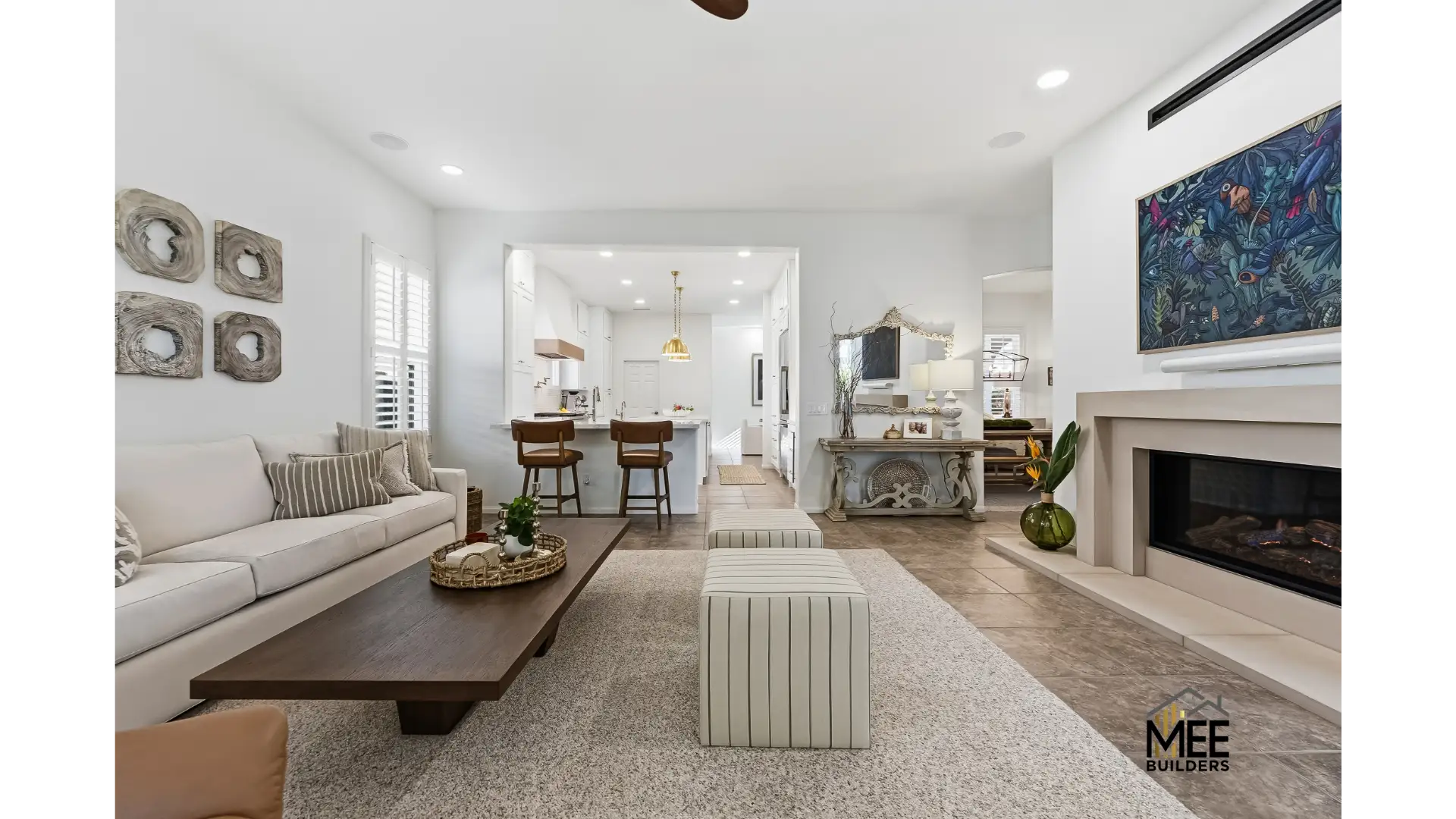 A modern open-concept living room featuring a neutral color palette with beige sofa, striped ottomans, and dark wood coffee table. The space flows into a white kitchen with bar seating, while decorative elements include a contemporary fireplace with colorful botanical artwork above it, wreath wall art, and a rustic console table. The room showcases clean lines, recessed lighting, and hardwood flooring throughout.