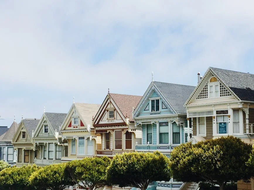A row of colorful Victorian-style houses known as the Painted Ladies, featuring ornate architectural details and distinctive peaked gables. The homes are painted in various pastel colors including cream, pink, blue, and yellow, with manicured green shrubs lining the front. These iconic San Francisco residences showcase classic Queen Anne architecture against a pale blue sky.