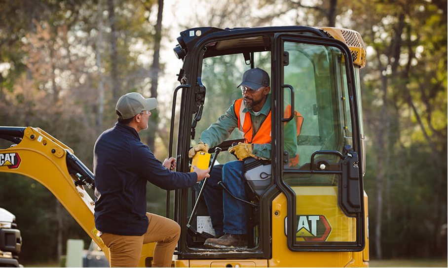 Two workers engage in conversation at a construction or work site, with one person standing on the ground handing a yellow thermos or beverage container to another person seated in the cab of a yellow CAT excavator. The operator wears an orange safety vest and cap, while the scene is set in a wooded area with trees visible in the background, suggesting an outdoor work environment during daytime.