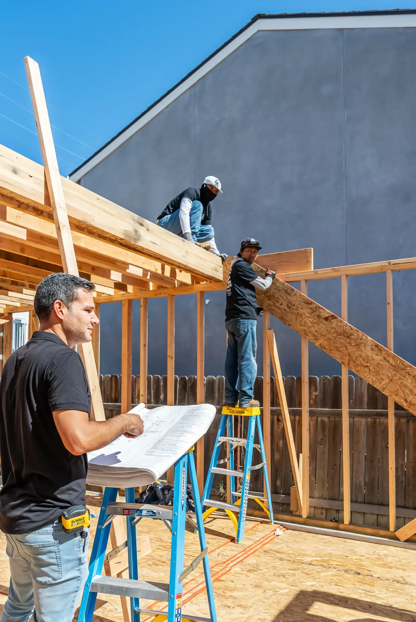 Three construction workers building a wooden frame structure at a residential construction site. In the foreground, a worker in a black shirt reviews blueprints while two others work on the upper level framing, with one standing on a yellow ladder. A gray modern building and clear blue sky are visible in the background.