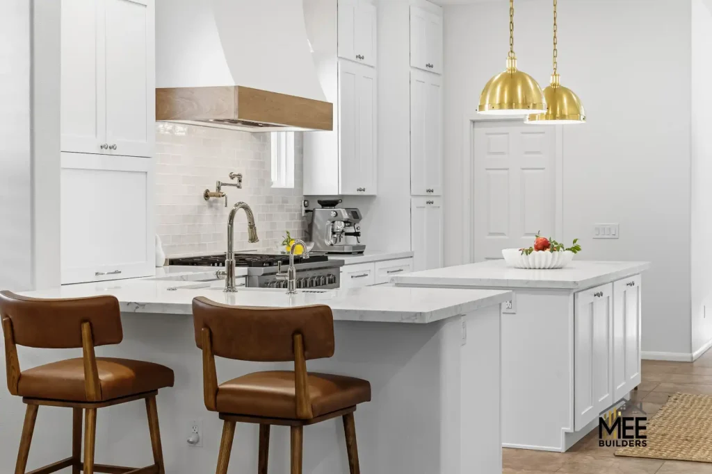 A modern white kitchen featuring a large island with marble countertops and two brown leather bar stools. The space includes white shaker-style cabinets, a professional-grade gas range with a wood-accented hood, and two brass pendant lights hanging over a secondary white island. The clean design incorporates subway tile backsplash and stainless steel appliances throughout.