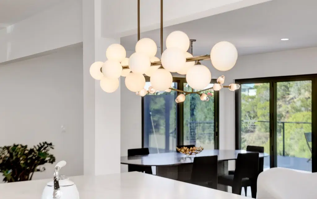 A modern dining room featuring a striking molecular-style chandelier with multiple glowing white globe lights suspended from brass or gold-toned arms. The space has floor-to-ceiling black-framed windows overlooking greenery, a sleek black dining table with matching chairs, and minimalist white walls. The contemporary lighting fixture serves as the room's focal point against the clean, monochromatic color scheme.