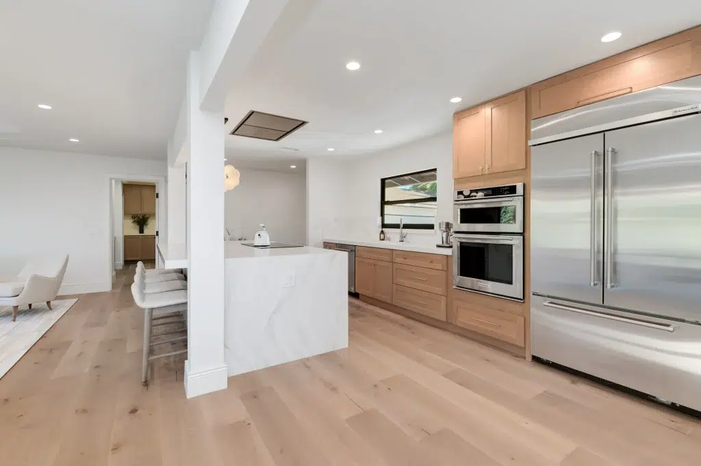 A modern open-concept kitchen featuring light wood cabinetry, stainless steel appliances including a built-in refrigerator and wall ovens, and a white waterfall-edge island with bar seating. The space showcases light hardwood flooring throughout, white walls, recessed lighting, and an open flow into adjacent living areas. The minimalist Scandinavian-inspired design creates a bright, airy atmosphere with clean lines and natural materials.