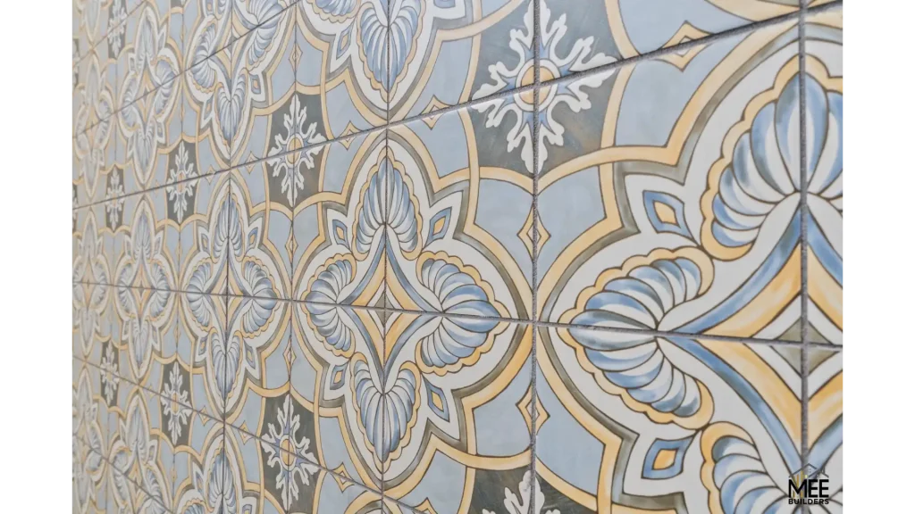 A decorative wall covered in ornate ceramic tiles featuring an intricate Moorish or Mediterranean-inspired pattern. The design showcases symmetrical floral and geometric motifs in soft blue, cream, and gold tones with detailed line work creating a luxurious, vintage aesthetic. The tiles appear to be hand-painted or printed with a traditional damask-style pattern that repeats across the surface.