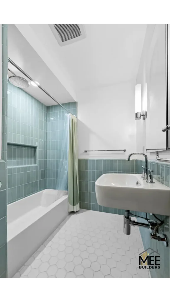 A modern bathroom featuring sage green vertical subway tiles on the walls, a white bathtub with chrome fixtures and rainfall showerhead, a wall-mounted rectangular sink, and white hexagonal floor tiles. The clean, minimalist design includes chrome grab bars, contemporary wall sconces, and a ceiling ventilation fan. The color scheme combines crisp white surfaces with calming seafoam green tile accents.