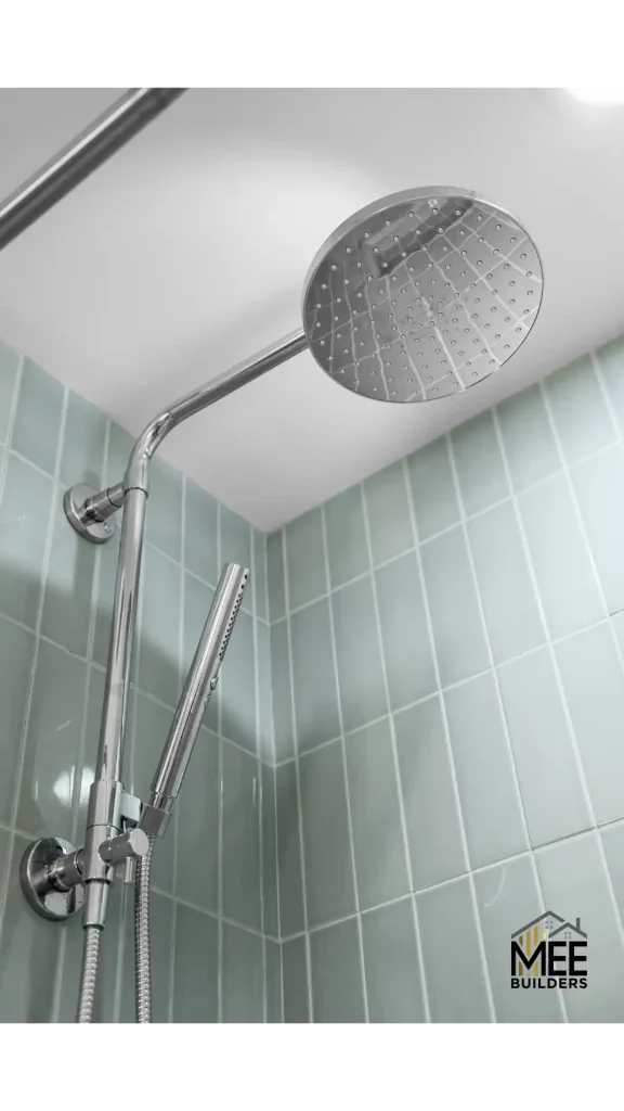 A chrome rainfall showerhead mounted on an adjustable arm extends from the corner of a bathroom with light blue-green vertical tile walls. The large circular showerhead features a perforated pattern for water distribution, and a separate handheld shower attachment with flexible hose is visible below. The modern fixture is photographed against a white ceiling in what appears to be a recently renovated shower enclosure.