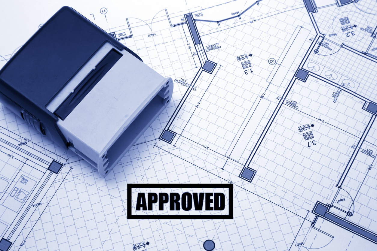 A rubber stamp and small architectural model house rest on top of detailed architectural blueprints showing room layouts and measurements. An "APPROVED" stamp impression in black text within a rectangular border is prominently displayed on the floor plan. The image represents the building permit approval process, with the blue-tinted technical drawings showing grid lines, room dimensions, and structural elements typical of construction documentation.
