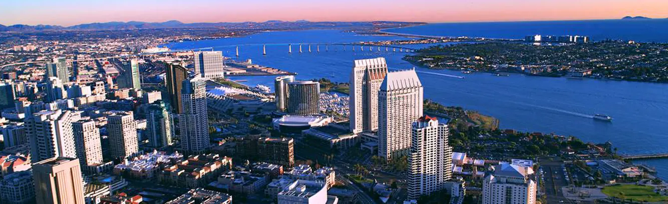 Aerial panoramic view of San Diego's downtown skyline and waterfront during sunset, featuring modern high-rise buildings, the San Diego Bay with its distinctive Coronado Bridge, and surrounding urban development. The image captures the city's coastal landscape with pink and blue twilight hues painting the sky, while sailboats dot the water and the distant hills frame the horizon.