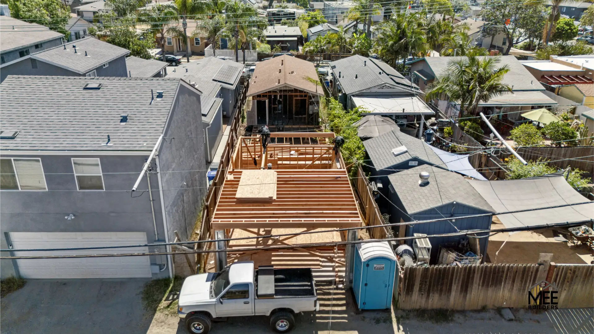 Aerial view of a residential construction site showing a narrow lot with exposed wooden framing for a new multi-level structure. The skeletal frame reveals floor joists and deck platforms under construction, with a white pickup truck and portable toilet visible at ground level. The property is tightly nestled between existing homes in a dense suburban neighborhood with mature trees.