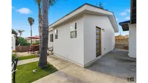 A modern white accessory dwelling unit (ADU) with a contemporary angled roofline sits in a residential backyard. The small structure features a wooden front door with glass panels, several windows, and sits on a concrete pad beside a palm tree. The surrounding yard includes artificial turf, outdoor furniture with a red umbrella, and wooden fencing, showcasing a typical Southern California residential setting.