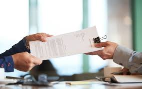 Two business professionals exchanging a contract or legal document across a desk, with one person in a dark suit handing papers to another person in a light-colored shirt who is holding a pen. The scene appears to be taking place in a bright office setting with windows visible in the background, suggesting a formal business transaction or agreement signing.