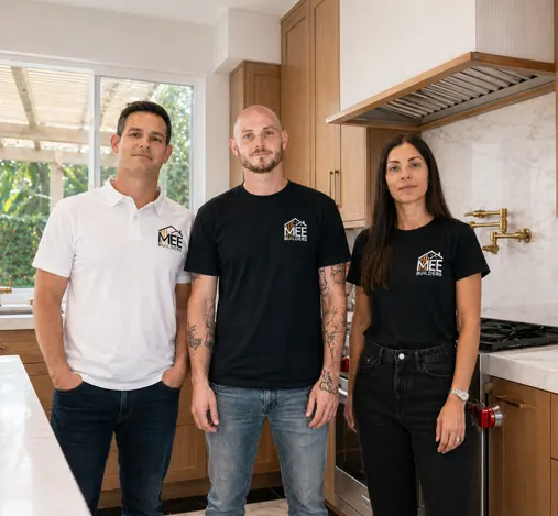 Three professionals from MBE Company stand in a modern kitchen with light wood cabinetry and marble backsplash. They wear matching company-branded shirts in white and black, positioned in front of a stainless steel range hood and large windows overlooking greenery. The team appears to be kitchen remodeling or home improvement specialists showcasing their work.