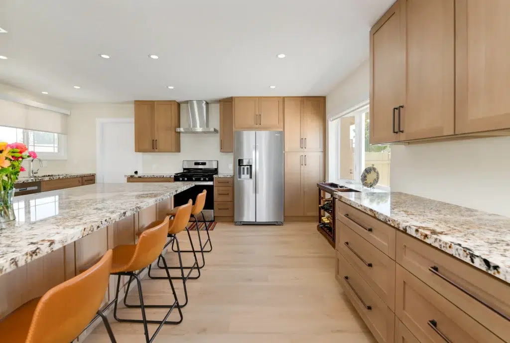 A modern kitchen featuring light wood cabinetry with black hardware, speckled granite countertops, and stainless steel appliances including a range hood and refrigerator. The space includes a kitchen island with three orange-tan leather bar stools with black metal frames. Light wood flooring and recessed lighting complete the bright, contemporary design with white walls and natural light from windows.