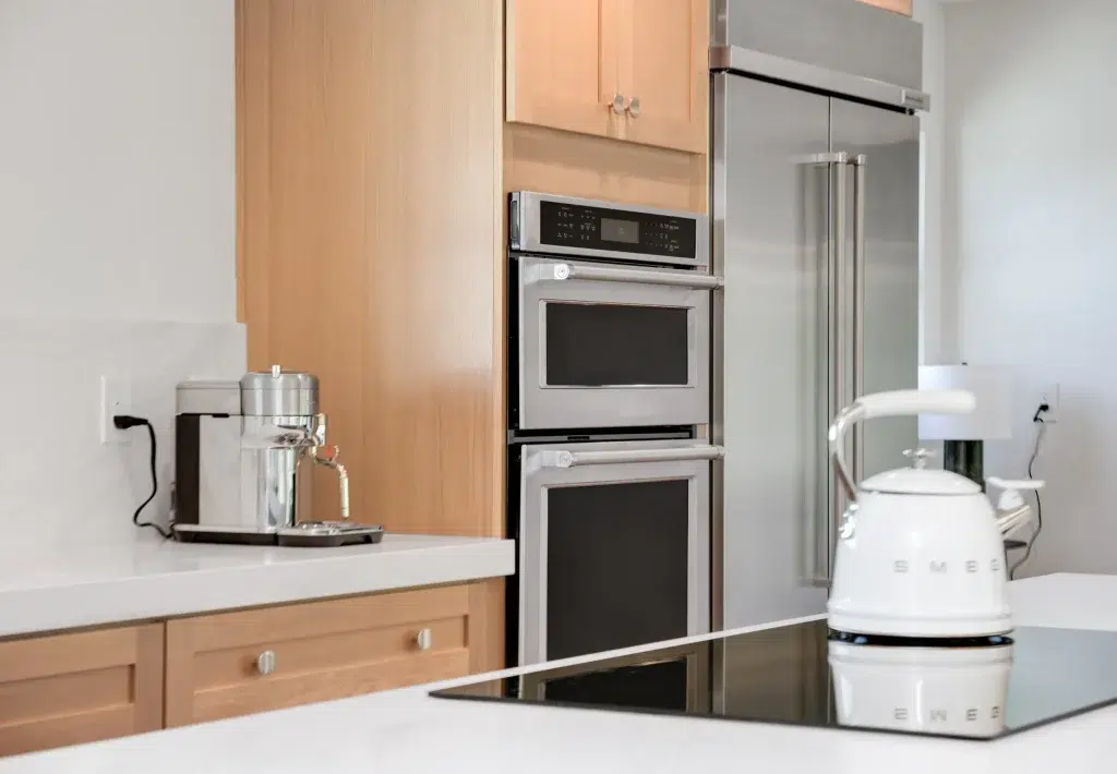 A modern kitchen featuring light wood cabinetry with stainless steel appliances, including a stacked double wall oven and microwave unit, a French door refrigerator, and a stovetop. White countertops display a retro-style white SMEG kettle and canister, along with a glass coffee maker on a tray. The kitchen has a clean, minimalist design with white walls and contemporary finishes.