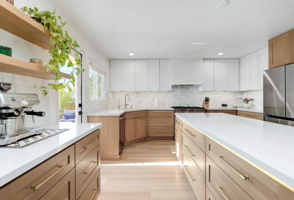 A bright modern kitchen featuring a two-tone design with natural wood lower cabinets and white upper cabinets. The space includes white quartz countertops, a white tile backsplash, stainless steel appliances, and light wood flooring. An espresso machine sits on the left counter beneath floating wooden shelves decorated with trailing plants, while a door opens to an outdoor area.