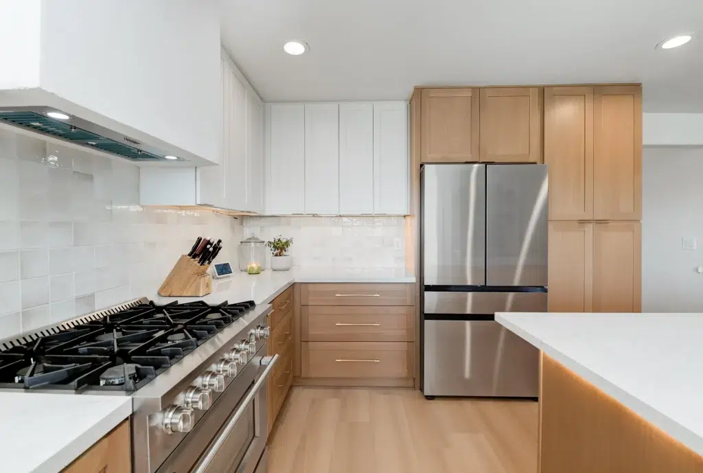 A modern L-shaped kitchen featuring a two-tone cabinet design with natural wood lower cabinets and white upper cabinets. The space includes white quartz countertops, a white subway tile backsplash, stainless steel appliances including a professional gas range and French-door refrigerator, and light wood flooring that complements the cabinetry.