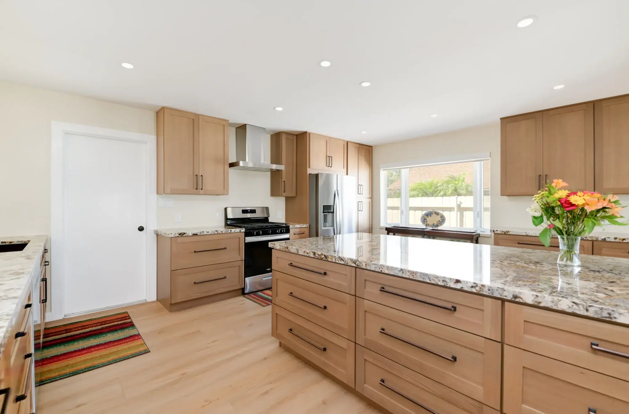 A modern kitchen featuring light wood cabinets with dark bronze hardware and speckled granite countertops. The space includes stainless steel appliances, a large window with a view of neighboring buildings, and light wood flooring complemented by colorful striped runners. Fresh flowers in a vase add a pop of color to the clean, contemporary design with white walls and recessed lighting.
