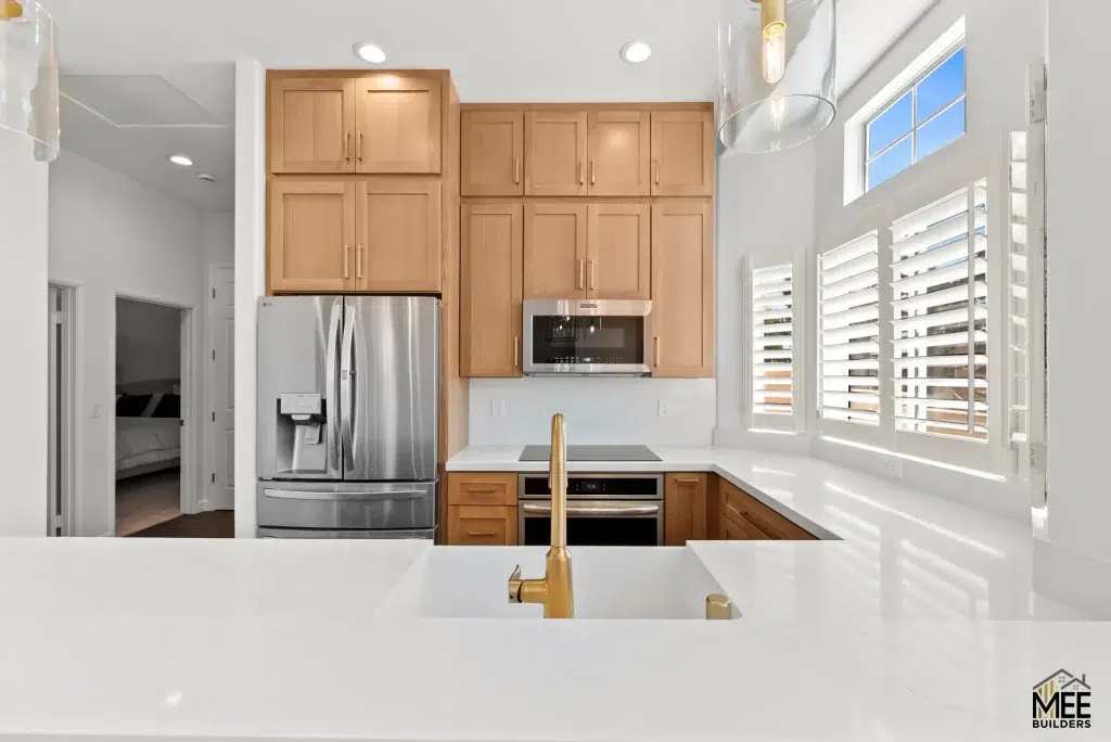 A modern kitchen featuring light natural wood cabinets arranged in a floor-to-ceiling configuration, complemented by white quartz countertops and a gold faucet visible in the foreground. The space includes stainless steel appliances including a French-door refrigerator and built-in microwave, with white plantation shutters on the windows allowing natural light to fill the room. The clean, contemporary design is accented by recessed lighting and opens to an adjacent hallway leading to a bedroom.