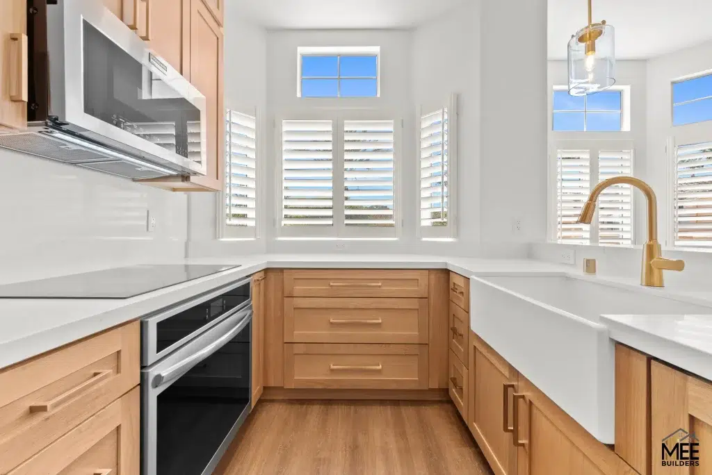A modern U-shaped kitchen featuring light wood Shaker-style cabinets paired with white quartz countertops and a white subway tile backsplash. The space includes stainless steel appliances, a brass faucet and pendant light fixture, and white plantation shutters on multiple windows that allow natural light to fill the room. The warm wood-tone flooring complements the natural wood cabinetry, creating a bright and contemporary cooking space.