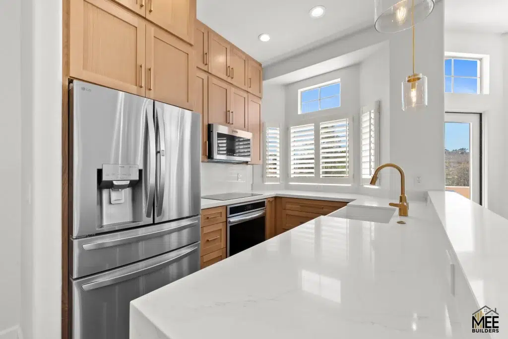 A modern kitchen featuring light wood shaker-style cabinets paired with white quartz countertops and a white island. The space includes stainless steel LG appliances, white plantation shutters on multiple windows, and a contemporary gold pendant light fixture. The bright, airy design showcases a clean aesthetic with recessed lighting and views of hills through the windows.