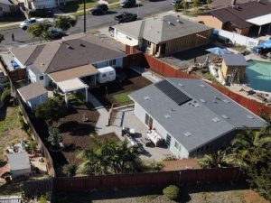 Aerial view of a residential neighborhood showing single-family homes with various roof types and yard spaces. The foreground features a white house with solar panels installed on its gray roof, surrounded by a landscaped backyard with palm trees and garden beds. Several other houses, cars parked on streets, and a mix of fencing and vegetation are visible throughout the suburban setting.