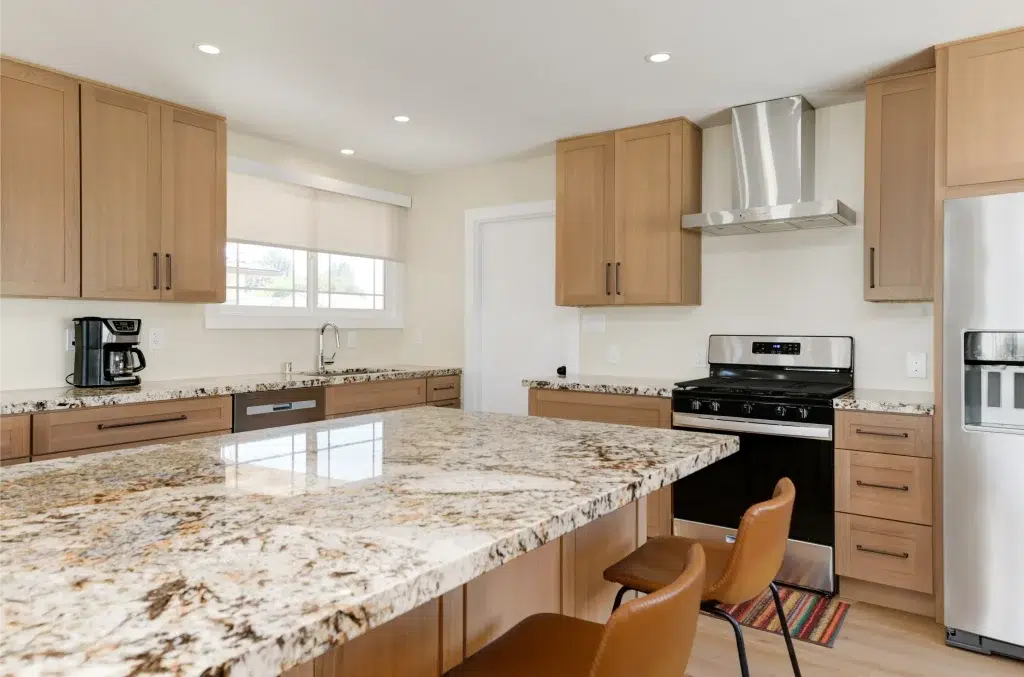 A modern kitchen featuring light wood cabinets with sleek black hardware, speckled granite countertops, and stainless steel appliances including a gas range and range hood. The space has a bright, airy feel with white walls, recessed lighting, and windows with white blinds. A kitchen island with matching granite takes center stage, accompanied by two tan-orange mid-century modern chairs and a colorful striped rug.
