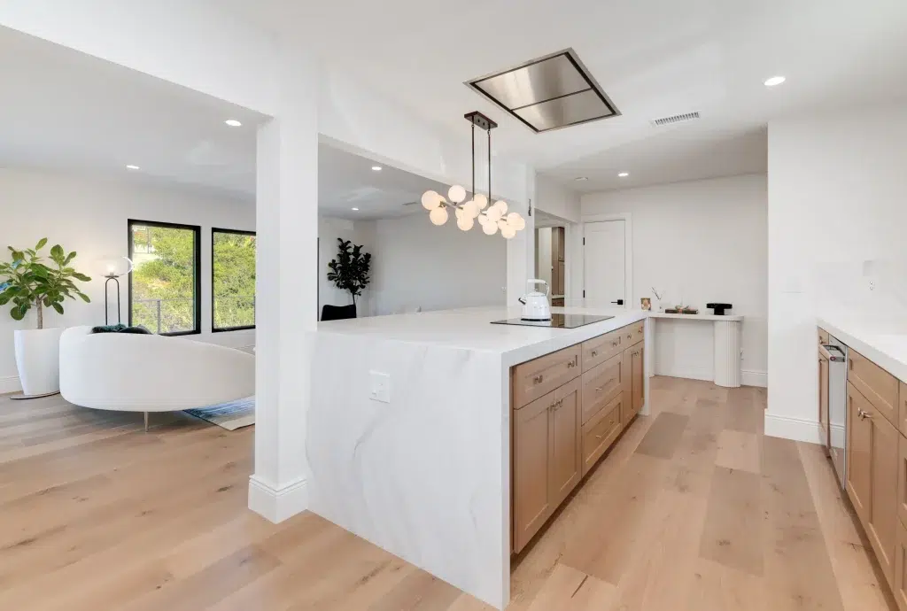 A modern open-concept kitchen featuring a large white marble waterfall island with natural wood cabinetry, white quartz countertops, and a contemporary globe pendant light fixture. The space flows into a bright living area with light hardwood flooring, white walls, and black-framed windows overlooking greenery. The design combines warm wood tones with crisp white finishes and includes a ceiling-mounted range hood above the island.
