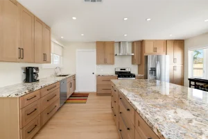 A modern kitchen featuring light wood cabinets with dark hardware and speckled granite countertops. The spacious layout includes a large central island, stainless steel appliances including a French door refrigerator and gas range with hood, and recessed lighting throughout. Light wood flooring and white walls create a bright, contemporary space with a colorful striped runner rug on the floor.