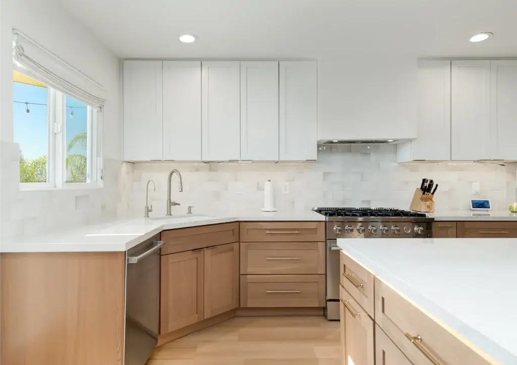 A modern two-tone kitchen featuring white upper cabinets and light wood lower cabinets with white quartz countertops. The space includes a professional-grade gas range, stainless steel appliances, and a white subway tile backsplash. Natural light streams through a window above the sink, which has a contemporary brushed nickel faucet, while the kitchen opens to what appears to be an island on the right side.