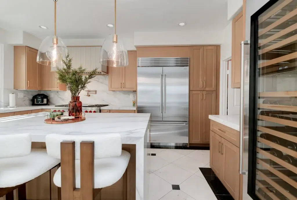 A modern kitchen featuring light wood cabinetry with a white marble countertop island. The space includes stainless steel appliances, including a built-in refrigerator and wine cooler, complemented by glass dome pendant lights with brass fixtures hanging above the island. White bar stools are positioned at the waterfall-edge island, and the backsplash displays a marble pattern matching the countertops.