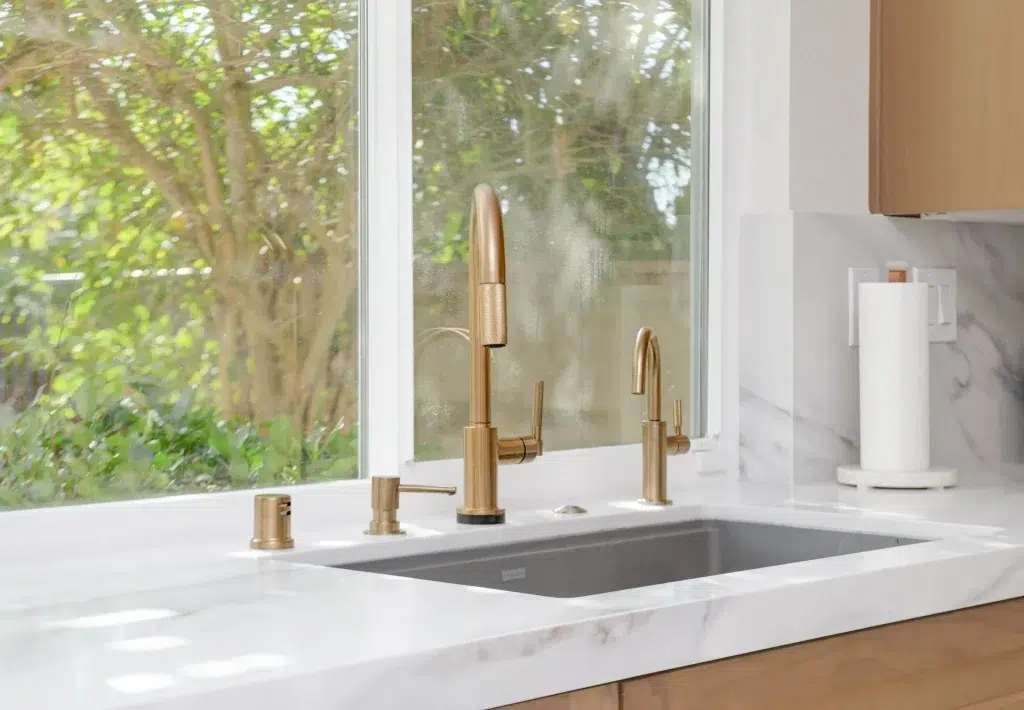 A modern kitchen sink features an elegant gold-toned articulating faucet with a pull-down spray head as its centerpiece, flanked by matching gold accessories including a soap dispenser and separate hot water tap. The sink is set in a white marble countertop with gray veining, positioned beneath a large window overlooking lush green foliage. Light wood cabinetry and white marble backsplash complement the contemporary design, with a white paper towel holder visible on the counter.