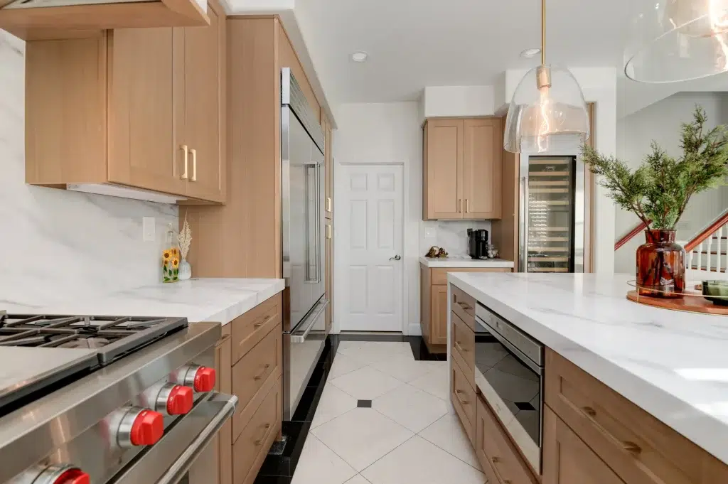 A modern kitchen featuring light wood cabinetry with white marble countertops and stainless steel appliances. The space includes a professional gas range with distinctive red knobs, built-in refrigerator, and wine cooler, with elegant glass pendant lighting hanging over a central island. The kitchen has white tile flooring and connects to other areas of the home through a white door visible in the background.