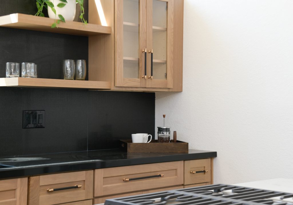 A modern kitchen featuring light oak cabinetry with glass-front upper cabinets and dark matte black countertops and backsplash. The open shelving displays glassware, while brass and black hardware accents the cabinet doors. A white coffee cup and French press sit on a wooden tray on the counter, with a gas cooktop visible in the foreground.