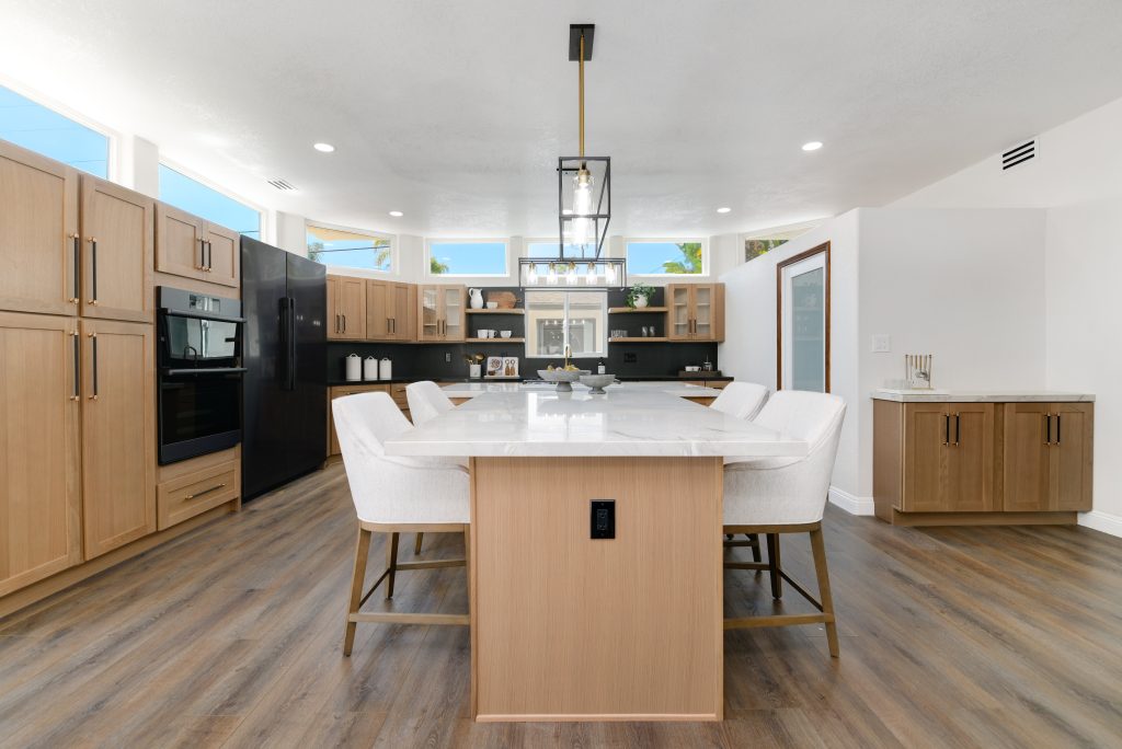 A modern open-concept kitchen featuring light wood cabinetry and a large white marble-topped island with four white upholstered bar stools. The space showcases black built-in appliances, a dark backsplash, and contemporary glass pendant lighting hanging from the ceiling. Natural light streams through clerestory windows above the cabinets, complementing the warm wood-toned flooring throughout.
