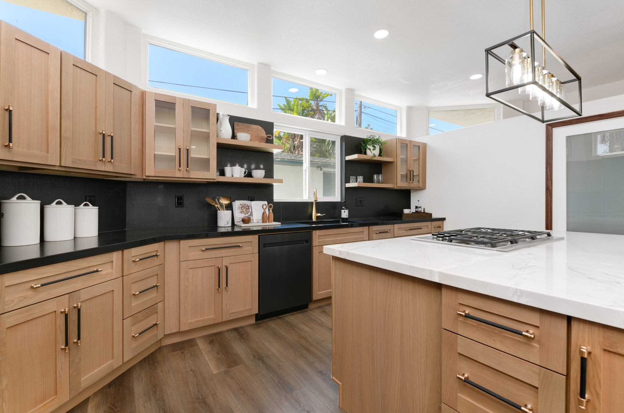 A modern kitchen featuring light wood cabinetry with black countertops along the walls and a white quartz waterfall island with built-in cooktop. The space includes black appliances, a geometric pendant light fixture, and a row of transom windows above the upper cabinets allowing natural light. The design combines warm wood tones with contemporary black and white elements throughout.