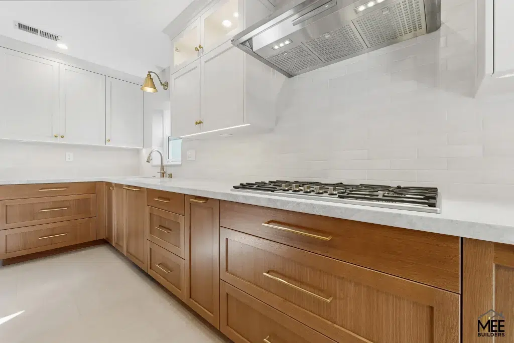A modern kitchen featuring two-tone cabinetry with natural wood lower cabinets and white upper cabinets, complemented by white quartz countertops. The space includes a gas cooktop, stainless steel range hood, white subway tile backsplash, and brass hardware and fixtures. The clean design showcases ample storage with multiple drawers in the wood-toned base cabinets and white tile flooring throughout.