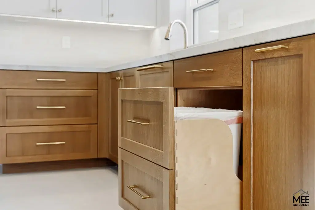 Modern kitchen featuring natural wood cabinets with brushed gold hardware and white quartz countertops. A pull-out trash bin drawer is shown open, revealing an integrated waste management system. The kitchen showcases clean lines with white upper cabinets and a white tiled backsplash.
