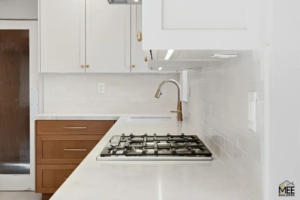 A modern L-shaped kitchen corner featuring white quartz countertops with a built-in gas cooktop and champagne gold faucet. The design combines white upper cabinets with warm wood-toned lower cabinets, complemented by a white subway tile backsplash. A stainless steel range hood is mounted above, and the space showcases a clean, contemporary aesthetic with mixed materials.