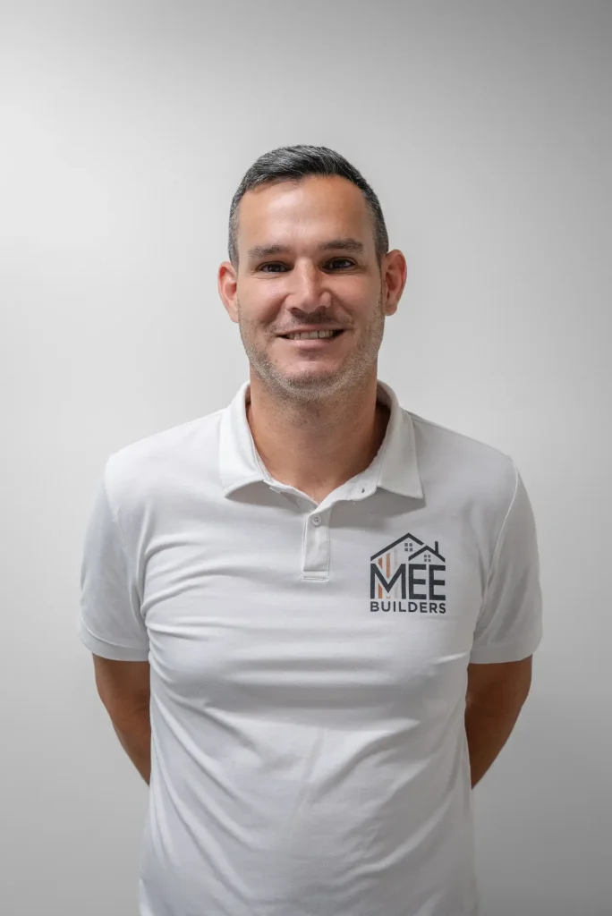 A professional headshot of a man wearing a white polo shirt with the MEE Builders logo featuring house icons. He has short dark hair with gray at the temples, a neatly trimmed beard, and is smiling warmly at the camera against a plain light gray background. His hands are positioned casually in his pockets in a relaxed, confident pose.