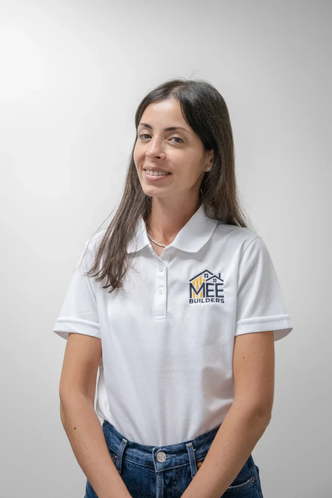 A woman with long dark hair wearing a white polo shirt with the MEE Builders logo embroidered on the chest stands against a plain light background. She is smiling warmly while looking slightly to the side, dressed professionally in the company uniform paired with dark blue jeans. The photo appears to be a professional headshot or team member portrait.