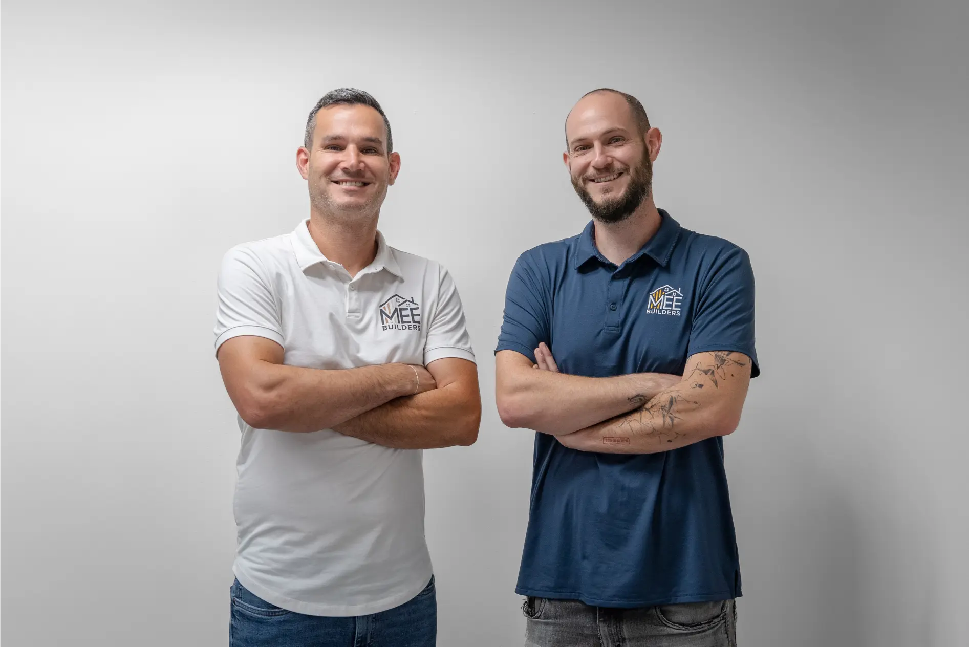 Two male construction professionals standing side by side with arms crossed, both wearing polo shirts embroidered with the MEE Builders logo. The man on the left wears a white polo while the man on the right wears a blue polo and has visible tattoos on his arm. Both are smiling confidently against a neutral gray background in a professional portrait setting.
