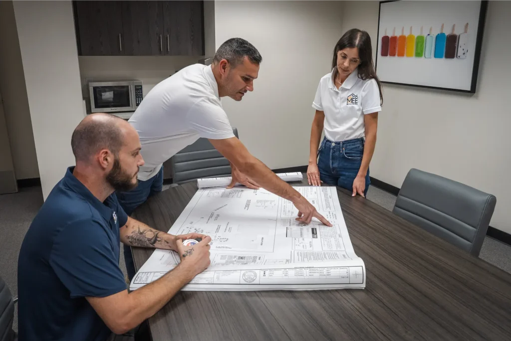 Three professionals reviewing architectural blueprints spread across a dark wood conference table in a modern office break room. A seated man in a navy polo shirt examines the plans while a man in white points to specific details, and a woman in a white MEE polo shirt observes. The contemporary space features dark cabinetry, a microwave, and colorful popsicle artwork on the wall.