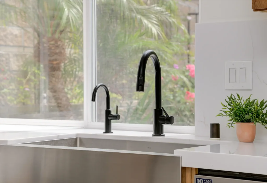 A modern kitchen sink with two sleek black faucets - a shorter beverage faucet on the left and a taller pull-down kitchen faucet on the right - set against a white countertop. Behind the sink is a large window overlooking palm trees and tropical landscaping, with a small potted plant in a terracotta pot visible on the counter to the right.