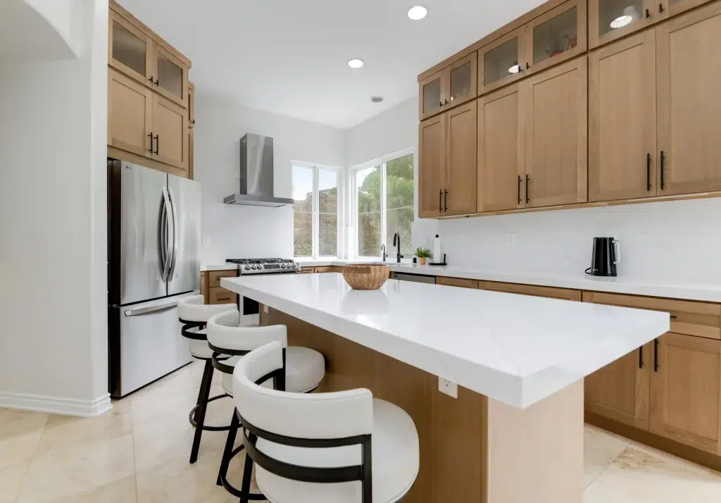 A modern kitchen featuring light wood cabinetry with black hardware, white quartz countertops, and a large island with three white upholstered bar stools. The space includes stainless steel appliances, recessed lighting, and windows providing natural light. The design combines warm wood tones with crisp white surfaces and black accents for a contemporary aesthetic.