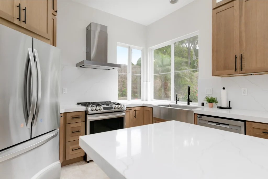 A modern kitchen featuring light wood cabinetry with black hardware, white quartz countertops, and stainless steel appliances including a French-door refrigerator and gas range with overhead vent hood. Large windows above the corner sink provide natural light and views of palm trees and greenery outside. The kitchen has a clean, contemporary design with a white island in the foreground and an open, airy layout with vaulted ceilings.