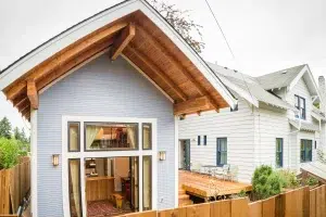 A modern two-story house featuring light gray horizontal siding with prominent exposed wood beam details under the peaked roof overhang. The front facade showcases large floor-to-ceiling windows with black frames arranged in a grid pattern, revealing a warm interior with wooden furnishings. A wooden deck extends from the side of the house, adjacent to a neighboring white residence, with landscaping visible in the foreground.