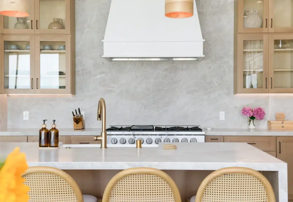 A modern kitchen featuring a white marble backsplash and light wood cabinets with glass-front upper cabinets. A professional-grade stainless steel range with a white hood vent serves as the focal point, flanked by a brass faucet at the kitchen island. Three cane-backed bar stools line the white countertop island in the foreground, with amber soap dispensers and pink peonies adding warm accents to the neutral color scheme.