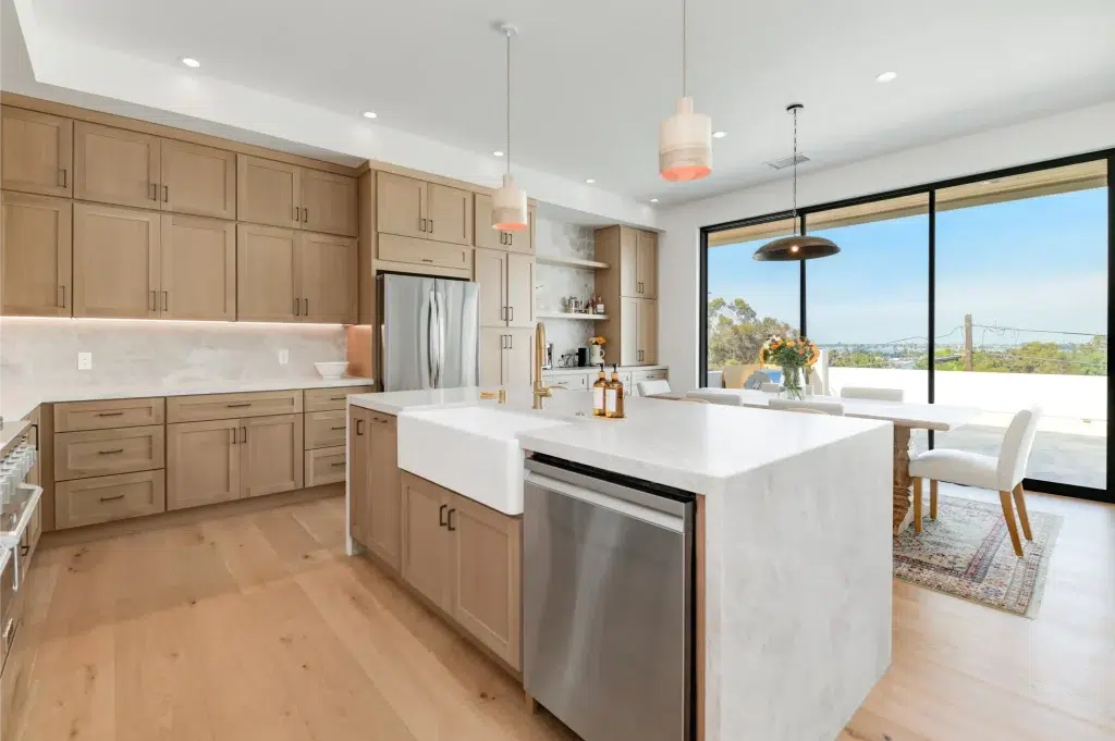 A modern kitchen featuring light wood cabinetry with shaker-style doors, a white marble waterfall island with an integrated farmhouse sink, and stainless steel appliances. The space opens to a dining area through large black-framed sliding glass doors that reveal scenic outdoor views. Pendant lights with coral-colored accents hang above the island, complementing the warm wood flooring that runs throughout.