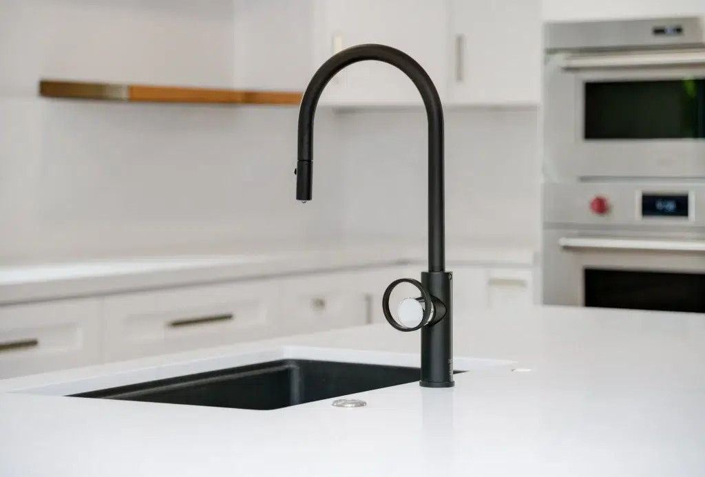 A modern matte black kitchen faucet with a curved gooseneck spout and circular side handle is mounted on a white countertop next to a black undermount sink. The faucet features a pull-down spray head and stands out against the minimalist white kitchen with built-in appliances visible in the background.